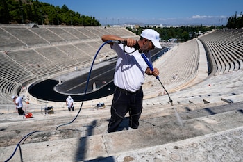 Hombre con gorra y camiseta blancas limpia gradas de un estadio con máquina de agua a presión. Se ven gradas vacías, una pista negra y otros trabajadores
