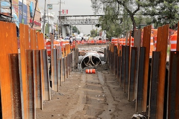 Sitio de construcción con pilotes metálicos, grandes tuberías blancas, conos de tráfico naranja, tierra y escombros, y un puente peatonal al fondo