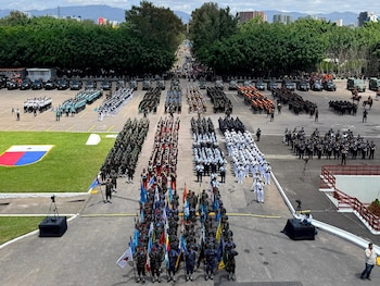 Grandes formaciones de militares con uniformes variados y banderas desfilan en una plaza asfaltada, junto a vehículos y un escudo en el césped