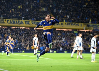 Un futbolista con uniforme azul y amarillo salta sobre el césped frente a una tribuna con público y otros jugadores