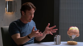 El Dr. David Eagleman, un hombre de cabello castaño y camisa azul, gesticula con ambas manos mientras habla. Un modelo de cerebro coloreado está a su derecha sobre una mesa