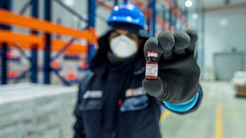 Persona con casco azul, mascarilla y guantes negros sostiene un pequeño vial de vacuna rosa en un almacén con estantes naranjas y azules. La vial tiene una etiqueta blanca