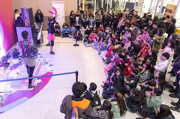 Dos artistas actúan en un escenario iluminado frente a un gran grupo de adultos y niños sentados en un suelo de baldosas blancas