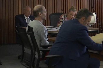 Un hombre con camiseta gris permanece sentado de perfil en una sala de tribunal junto a abogados y personal judicial