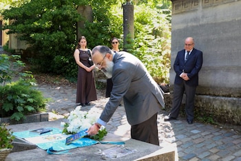 Hombre barbudo agachado coloca libro sobre ofrenda floral blanca y tela azul/blanca en tumba. Tres personas observan de pie. Entorno de jardín con árboles y piedra