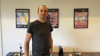 Hombre sonriendo ligeramente, vestido con una camiseta oscura, de pie frente a una pared clara con tres pósters enmarcados y un escritorio con una cafetera y una taza