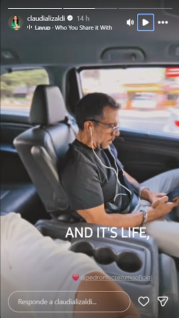 Un hombre de mediana edad con barba y gafas sentado en el asiento trasero de un coche oscuro, usando auriculares blancos y mirando su teléfono móvil