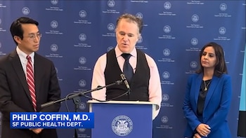 Philip Coffin, MD, do Departamento de Saúde Pública de São Francisco, fala no palco, junto com um homem e uma mulher. Camisa azul com logotipo