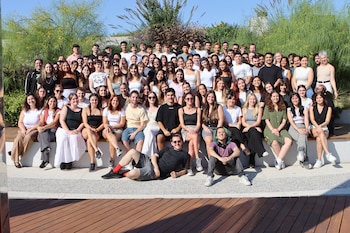 Un grupo grande y diverso de jóvenes posa al aire libre. Algunos de pie, otros sentados en un banco y en el suelo, frente a vegetación y un cielo azul