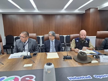 Tres hombres sentados en una mesa de conferencia con micrófonos, documentos y un sombrero. Uno de ellos usa un chaleco con el logo de 'Procuraduría'