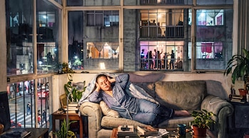 Ilustración en acuarela de una mujer en un sofá tapándose los oídos frente a un ventanal con vistas a una fiesta en otro edificio.