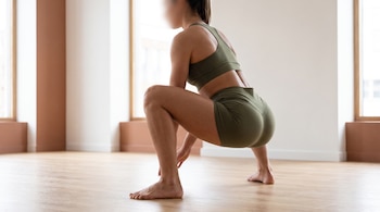 Una mujer vestida con ropa deportiva verde oliva realiza una sentadilla profunda descalza sobre un piso de madera.