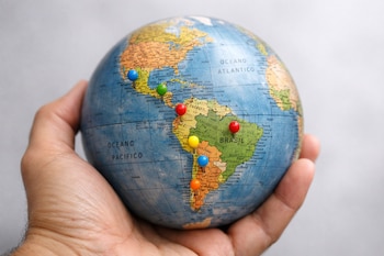 Mano adulta sostiene un globo terráqueo que muestra América Latina, con seis pines de colores (azul, verde, rojo, amarillo, celeste, terracota) en varios países.