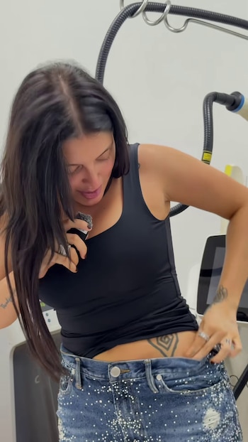 Una mujer de cabello oscuro baja la pretina de su pantalón para mostrar un tatuaje en la cadera junto a un equipo médico