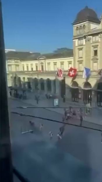 Vista aérea de una calle suiza con edificios de piedra clara y arcos en la planta baja, gente caminando, y banderas suizas en el balcón bajo un cielo azul