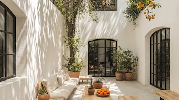 Patio exterior soleado con paredes blancas, grandes ventanales negros, un sofá largo beige, mesas de madera y plantas en macetas, incluyendo naranjas en un tazón.