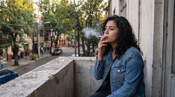 Mujer joven con cabello rizado oscuro y chaqueta de mezclilla fuma un cigarrillo en un balcón. Al fondo, una calle urbana con árboles y vehículos.