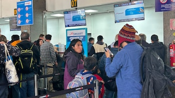 Grupo de pasajeros con mochilas y equipaje hace fila en mostradores de check-in del aeropuerto con pantallas de información de vuelos y un extintor rojo