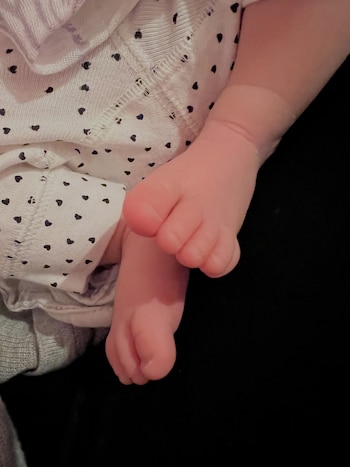 Dos pies de bebé con piel rosada asoman de una prenda blanca con diseño de pequeños corazones negros sobre un fondo oscuro