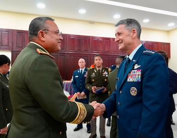 Primer plano de dos oficiales militares de diferentes ramas uniformados, uno de verde y otro de azul, estrechándose las manos y sonriendo
