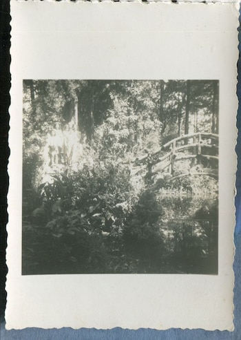 Fotografía monocromática de un grupo de personas de pie entre vegetación densa, con un puente rústico de madera visible en el fondo a la derecha