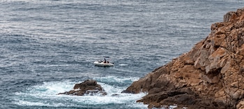 Una pequeña embarcación con tres personas a bordo en el mar, cerca de una costa rocosa en un día nublado. El agua presenta olas pequeñas