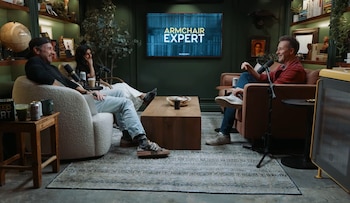Tres personas sentadas en sillones con micrófonos frente a una mesa de café. Una pantalla al fondo muestra el logo Armchair Expert. Estanterías con libros y plantas