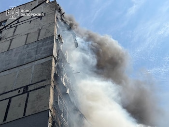 Fachada de un edificio gris de varios pisos con daños extensos y ventanas rotas, de donde sale abundante humo oscuro. Cielo azul en el fondo
