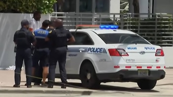 Vista trasera de un vehículo policial Ford Interceptor blanco de Miami con luces azules encendidas, y varios oficiales de policía de pie junto a un individuo