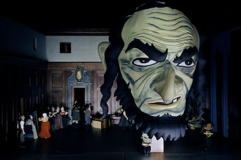 Escenario de ópera con una cabeza inflable gigante de un personaje, un grupo de actores, utilería, una puerta y paredes que simulan una sala