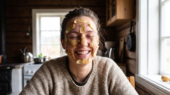 Persona sonriendo con cáscaras de papa cubriendo su rostro, vestida con un suéter de lana, sentada en una cocina rústica con paredes de madera y una ventana.