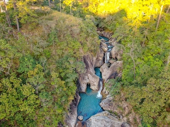 Vista aérea de cascadas y pozas de agua turquesa entre rocas y vegetación verde. Se aprecian cables de tirolina y luz dorada en la cima de los árboles