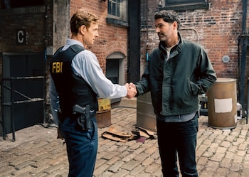 Nick Gehlfuss con chaleco negro del FBI y Tom Ellis con chaqueta oscura se dan la mano en un callejón de adoquines, con paredes de ladrillo al fondo