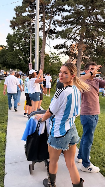 Una mujer rubia con camiseta de Argentina, shorts de jean y botas, empuja un cochecito con bandera. Tiene pintura azul en la mejilla y sonríe. Hay gente detrás