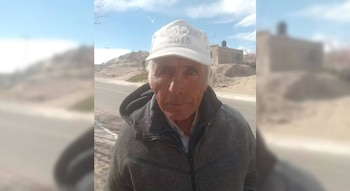Un hombre mayor con gorra blanca y chaqueta gris de pie junto a una carretera en un entorno árido