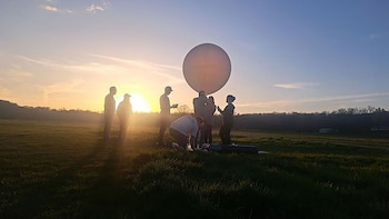 Grupo de personas en silueta en un campo de hierba, preparando un gran globo blanco. El sol bajo en el horizonte proyecta largas sombras