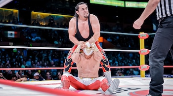 Un luchador de pie sujetando por la cabeza a un luchador enmascarado sentado en la lona de un cuadrilátero, junto a un árbitro.