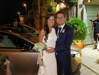 Maje Moreno Cantó y un hombre sonríen, vestidos de novia y novio, de pie frente a un coche. Se ve un ramo, vegetación y luces nocturnas