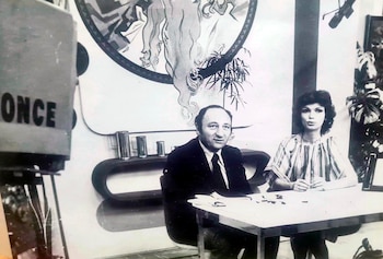 Un hombre y una mujer sentados en una mesa, con una cámara de televisión que tiene la palabra "ONCE" y un mural de fondo con plantas