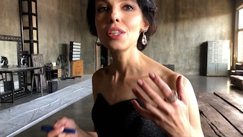 Primer plano de Julia Loktev, una mujer de cabello oscuro, aretes llamativos y vestido negro, gesticulando en un estudio con paredes de cemento