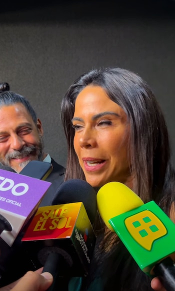 Uma mulher de cabelos escuros fala em vários microfones. Homem barbudo sorrindo levemente ao fundo