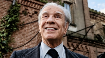 Jorge Luis Borges viste traje y corbata con la mirada en alto frente a un edificio antiguo de ladrillos cubierto de vegetación.
