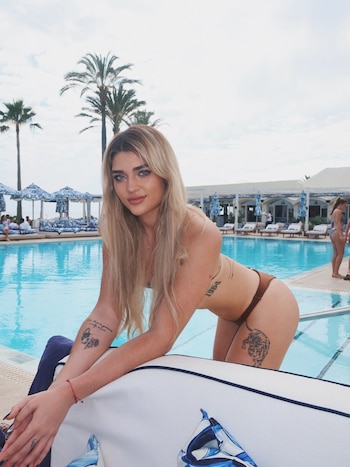 Marta Fort, una mujer rubia con ojos azules, posa en bikini marrón junto a una piscina. Se ven palmeras y sombrillas azules y blancas al fondo