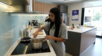 Daniela Celis, con delantal a rayas, cocina en una olla sobre una estufa eléctrica moderna en una cocina luminosa. Hay otros elementos de cocina visibles y personas al fondo.