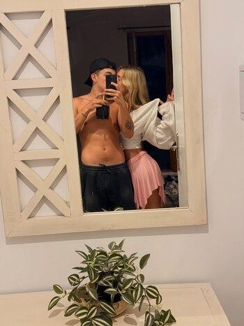 Reflejo en un espejo de una pareja besándose; el hombre, sin camiseta y gorra, sostiene un teléfono, y la mujer, de rosa, apoya su brazo