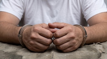 Primer plano de dos manos masculinas esposadas, entrelazadas sobre una mesa cubierta con tela de saco, con una persona en camiseta blanca.