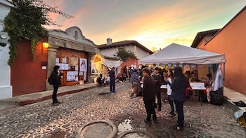 Personas se reúnen en una calle adoquinada con edificios históricos y pancartas; una carpa blanca y un cielo de atardecer son visibles