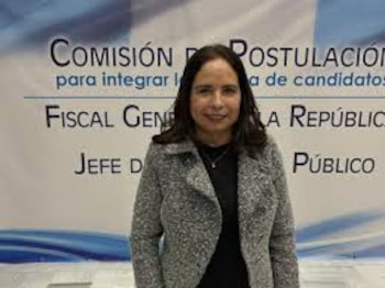 Mujer con cabello oscuro y abrigo gris de pie frente a un banner azul y blanco que dice 'Comisión de Postulación' y 'Fiscal General de la República'