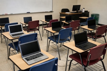 Salón de clases vacío con escritorios de madera, sillas azules y rojas, y laptops abiertas en cada escritorio. Una pizarra blanca y un escritorio de profesor.