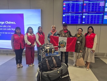 Varias personas en un aeropuerto, algunas con chalecos rojos y otras con una bandera de Perú, junto a un carrito con equipaje y pantallas de información de vuelos